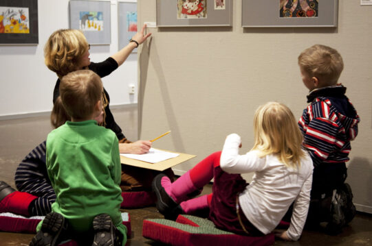 Neljä lasta ja aikuinen katsovat yhdessä seinällä olevaa taideteosta ja lukevat teostietoja taulun vieressä olevalta teoskortilta.